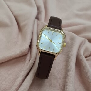Puede incluir: Un reloj de pulsera de forma cuadrada con una correa de cuero marr&oacute;n y una caja de color dorado. La esfera del reloj es blanca con marcadores de hora y manecillas de color dorado.