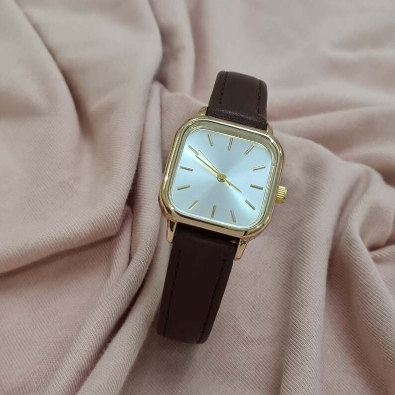 Puede incluir: Un reloj de pulsera de forma cuadrada con una correa de cuero marr&oacute;n y una caja de color dorado. La esfera del reloj es blanca con marcadores de hora y manecillas de color dorado.