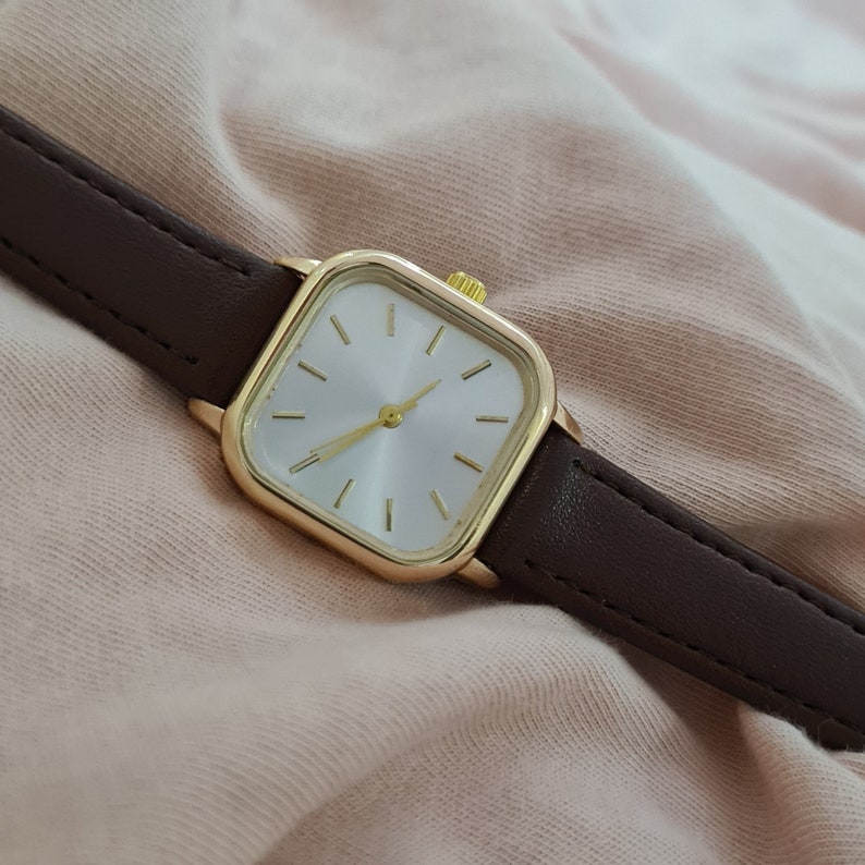 Puede incluir: Un reloj cuadrado dorado con una esfera blanca y una correa de cuero marr&oacute;n.