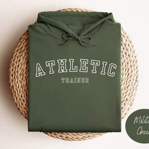 Könnte beinhalten: Ein grüner Kapuzenpullover mit dem Text "ATHLETIC TRAINER" in Weiß.