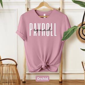 Peut inclure: Un t-shirt rose avec le texte "Payroll Squad" en lettres blanches. Le t-shirt est sur un cintre en bois et a un bas noué.