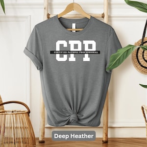 Può includere: Una maglietta grigio scuro con la scritta "CPP" in grandi lettere bianche sopra le parole "CERTIFIED PAYROLL PROFESSIONAL" in lettere bianche più piccole. La maglietta è annodata in basso.