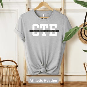 Könnte beinhalten: Graues T-Shirt mit dem Text "CTE" in weißen Buchstaben und den Worten "Career Technical Education" in schwarzen Buchstaben darunter. Das Shirt hängt an einem Kleiderbügel und hat einen geknoteten Saum. Der Text "Athletic Heather" befindet sich unter dem Shirt.