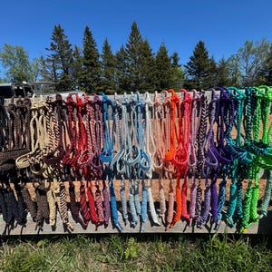 May include: A collection of colorful horse halters hanging on a wooden fence. The halters are displayed in a rainbow of colors, including black, brown, red, blue, orange, purple, and green. The sky is blue with trees in the background.