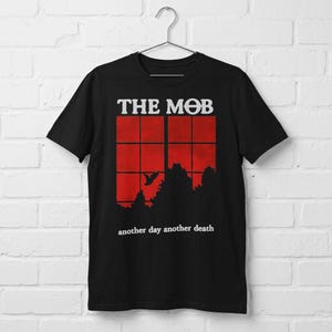Peut inclure: Un t-shirt noir à manches courtes avec une impression graphique portant le texte THE MOB en haut et another day another death en bas. Le motif central représente un cadre de fenêtre rouge à carreaux, une silhouette d'oiseau et des arbres.
