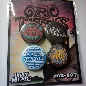 May include: A pack of four Ozric Tentacles badges. The badges display the band's logo in different colours and styles, including teal, black, blue, and yellow. The badges are packaged on a card with the Roxy Music logo and the text "POX-107".