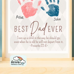 May include: A printable art print with a grey and white diamond pattern background. The print features two handprints, one pink and one blue, with the names Allie and Jake written below. The text "BEST Dad EVER" is written in brown lettering. A Bible verse, "Train up a child in the way he should go; even when he is old he will not depart from it. &bull; Proverbs 22:6.", is written below the handprints. The print has a white cloud with a rainbow and the text "Leru's PRINTABLES STUDIO" in the bottom left corner.