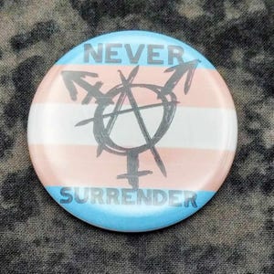 Könnte beinhalten: Ein runder Button mit einem Design der Transgender-Pride-Flagge. Der Button hat einen hellblau-rosa gestreiften Hintergrund mit den Wörtern "NEVER" und "SURRENDER" in Dunkelgrau. Ein schwarzes Symbol befindet sich in der Mitte.