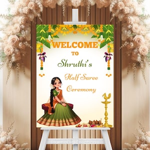 Può includere: Un cartello di benvenuto per una cerimonia Half Saree. Il cartello presenta il testo "WELCOME TO Shruti's Half Saree Ceremony". C'è un'illustrazione di una donna con un sari. Il cartello è su un cavalletto bianco.