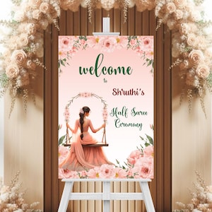 May include: A welcome sign for a Half Saree Ceremony. The sign features a floral design with the words "welcome to Shruti's Half Saree Ceremony". The sign is on a white easel, with floral decorations in the background.