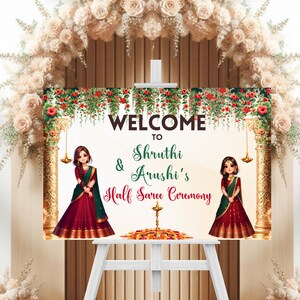 May include: Welcome to Shruthi & Arushi's Half Saree Ceremony sign with two girls in traditional Indian attire. The sign is decorated with flowers and gold accents.