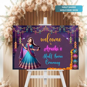 May include: Purple welcome sign with gold lettering for a half saree ceremony. The sign features a cartoon illustration of a woman wearing a blue and purple saree, with the text "Welcome to Arushi's Half Saree Ceremony".