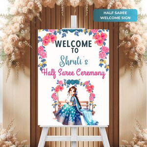 May include: A welcome sign for a Half Saree Ceremony. The sign displays the text "WELCOME TO Shruti's Half Saree Ceremony" with floral and illustration accents. The sign is on a white easel, with a floral arch in the background.