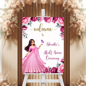 May include: A sign for "Shruthi's Half Saree Ceremony" with a cartoon illustration of a woman in a pink saree. The sign is decorated with pink flowers and the words "welcome to" in gold script.
