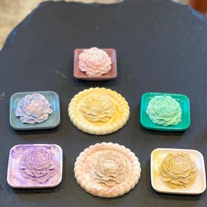 May include: An assortment of decorative floral-shaped soaps in various colors, including pink, yellow, green, and purple. The soaps are displayed on small square and round dishes, creating a visually appealing arrangement.