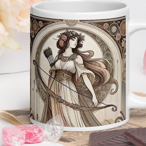 May include: White ceramic mug with a brown and beige illustration of a woman with long hair wearing a crown and holding a bow and arrow. The illustration is surrounded by a decorative frame.
