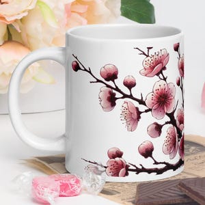 May include: White ceramic mug with a pink and white floral design. The design features a branch of cherry blossoms with pink flowers and green leaves.