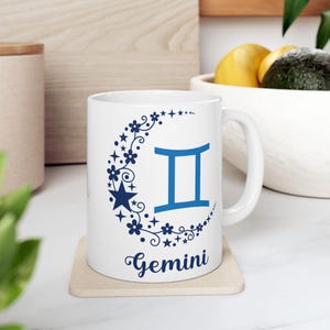 Op de afbeelding: Witte keramische mok met een blauw halve maan ontwerp met een bloemmotief en het sterrenbeeld Tweelingen. De mok heeft ook het woord "Tweelingen" in blauw onder het ontwerp.
