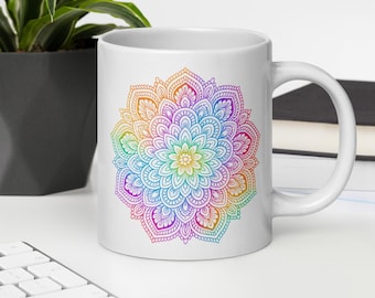 May include: White ceramic mug featuring a vibrant, multi-colored mandala design. The mandala showcases a spectrum of colors, including pink, orange, yellow, green, and blue, arranged in a symmetrical pattern. The mug has a curved handle and is set on a white surface.