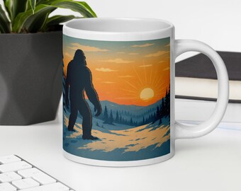 May include: White ceramic mug featuring a Bigfoot illustration in a snowy landscape with a sunset. The design includes a silhouette of the creature, mountains, and pine trees, with orange, blue, and white colors.