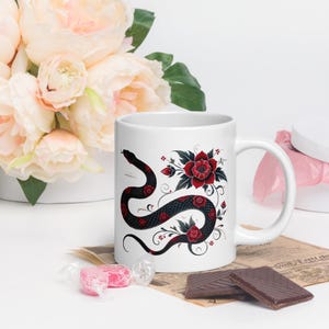 May include: White ceramic mug with a black snake wrapped around red roses and green leaves. The snake is curled around the roses and the design is on one side of the mug.