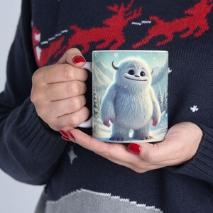 May include: White ceramic mug featuring a colourful illustration of a white yeti standing in a snowy landscape. The yeti has a friendly expression and is wearing a red scarf.