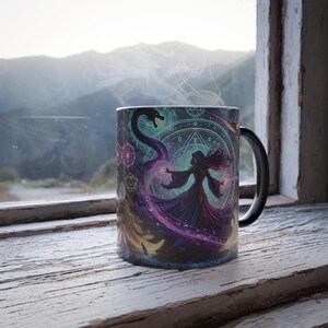 May include: A black ceramic mug with a colorful design featuring a woman in a purple robe casting a spell with a dragon. The design includes geometric shapes and symbols.