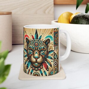 May include: A white ceramic mug with a colorful illustration of a jaguar's face with geometric patterns and feathers. The background is a gold and brown color with geometric patterns.