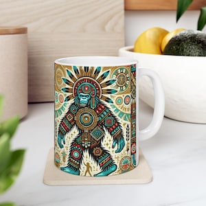 May include: A white ceramic mug featuring a colourful graphic of a mythical creature wearing a headdress. The creature is surrounded by geometric patterns and symbols. The mug is on a beige coaster.