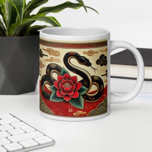May include: White ceramic mug with a black snake and red flower design. The snake is coiled around the flower and has a stylized, traditional Japanese look. The background is a cream color with red, gold, and black accents.
