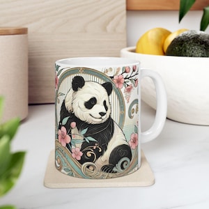 May include: White ceramic mug with a black and white panda bear illustration surrounded by pink flowers and gold accents. The mug has a handle and is sitting on a beige coaster.