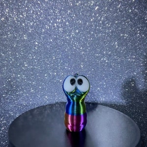 May include: A colorful, 3D printed, worm-like figure with large eyes, standing on a black surface against a glittery, silver background. The figure is rainbow colored and has a whimsical, cartoonish appearance.