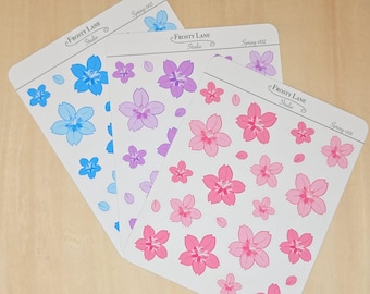 Pode incluir: Três folhas de autocolantes retangulares com a etiqueta Frosty Lane Studio e Spring 001, 002 e 003. Cada folha apresenta ilustrações de flores de cinco pétalas de vários tamanhos em azul, roxo e rosa, com pequenas pétalas espalhadas no fundo branco.
