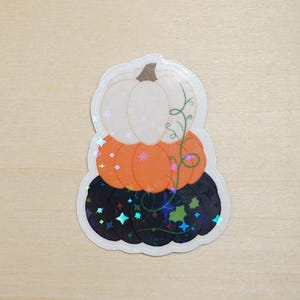 Könnte beinhalten: Ein Aufkleber mit einem Stapel von drei Kürbissen. Der oberste Kürbis ist weiß, der mittlere Kürbis ist orange und der unterste Kürbis ist schwarz mit einem glitzernden Finish. Die Kürbisse haben eine Ranken mit Blättern.