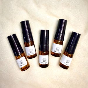 May include: Five small, brown glass bottles with black caps are arranged on a cream-colored surface. Each bottle has a white label with a heart and sun design and the words "Divine Love". The bottles are filled with a golden liquid.