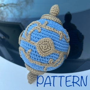 May include: A crocheted ornament in the shape of a blue and gold globe with a pattern of leaves and vines. The ornament has a gold top and is sitting on a car's windshield. The word "PATTERN" is written in blue letters below the ornament.