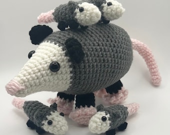 May include: A crocheted grey and white opossum with pink accents and three smaller crocheted opossums. The larger opossum is sitting with its tail curled around its body. The smaller opossums are sitting on top of the larger opossum.