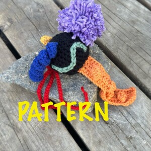 May include: A crocheted toy with a purple pom-pom head, black body, blue and green details, and orange and red legs. The toy is sitting on a piece of wood with the word "PATTERN" written in yellow letters.