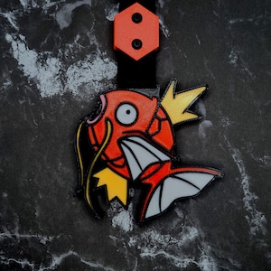 Può includere: Un portachiavi stampato in 3D raffigurante un pesce dei cartoni animati rosso e bianco con dettagli gialli. Il portachiavi ha un cinturino nero e un elemento superiore esagonale arancione con due cerchi neri. Il pesce ha un occhio grande e pinne dettagliate.