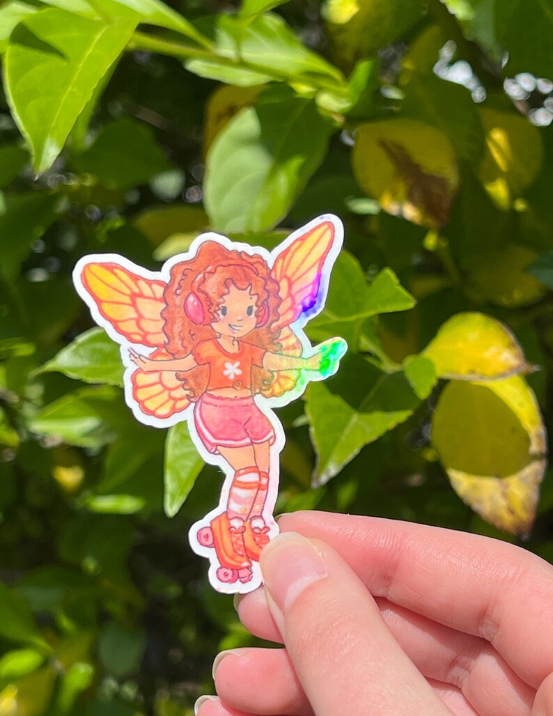May include: A vibrant sticker of a fairy on roller skates. The fairy has orange hair, headphones, and iridescent wings. She wears a pink outfit with orange accents. The sticker is held against a backdrop of green leaves.