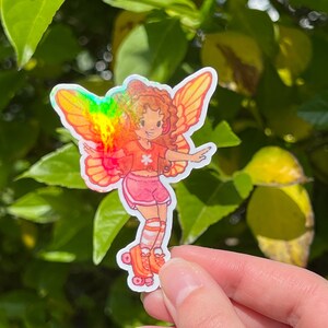 May include: A sticker depicting a cartoon fairy with orange hair, wearing a red top, pink shorts, and roller skates. The fairy has rainbow-coloured wings. The background is a blur of green leaves.