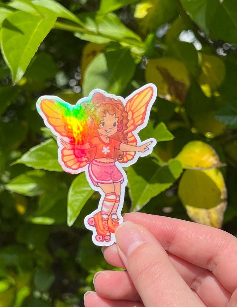 May include: A sticker depicting a cartoon fairy with orange hair, wearing a red top, pink shorts, and roller skates. The fairy has rainbow-coloured wings. The background is a blur of green leaves.