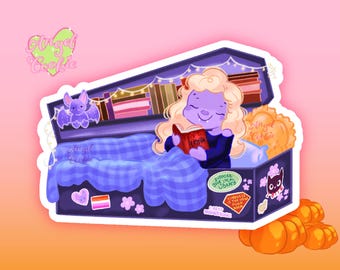 May include: A sticker illustration of a purple-skinned character with blonde hair reading a book inside a dark coffin. The coffin features a blue plaid blanket, a bat, bookshelves, and stickers that read Support Your Local Library and I Read Banned Books.