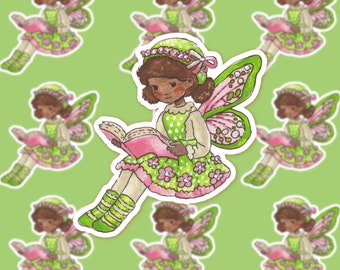 May include: A sticker featuring a cartoon fairy with brown skin, green and pink wings, and a green dress with white polka dots. She is reading a pink book and has a green hat with a pink bow.