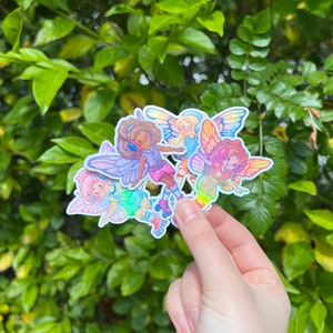 May include: A cluster of vibrant stickers showcasing cartoon fairies with butterfly wings. The fairies are in different poses, some wearing roller skates and headphones. The stickers have a holographic, iridescent finish, and are held against a backdrop of green leaves.