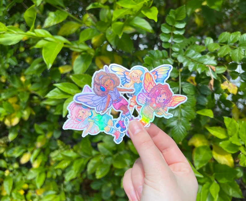 May include: A cluster of vibrant stickers showcasing cartoon fairies with butterfly wings. The fairies are in different poses, some wearing roller skates and headphones. The stickers have a holographic, iridescent finish, and are held against a backdrop of green leaves.