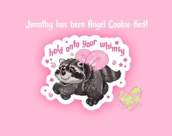 May include: A sticker featuring a grey and black raccoon with pink wings, surrounded by stars and swirls. The text reads "Jimothy has been Angel Cookie-fied!", "hold onto your whimsy", and "Angel Cookie" on a light green heart shape.