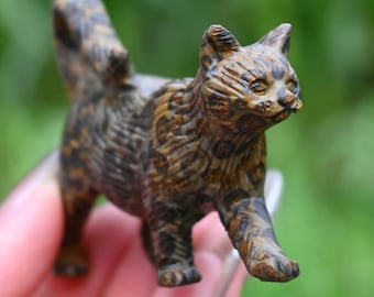 May include: A small, detailed figurine of a cat, carved from a brown and tan material. The cat is depicted in a walking pose, with its tail raised. The figurine has textured details, giving it a realistic appearance.