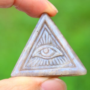 May include: A triangular shaped, carved stone pendant with an all-seeing eye design. The eye is carved in detail and has a light gray color.