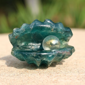 Puede incluir: Una concha de vidrio azul verdoso con una perla de vidrio transparente en su interior.